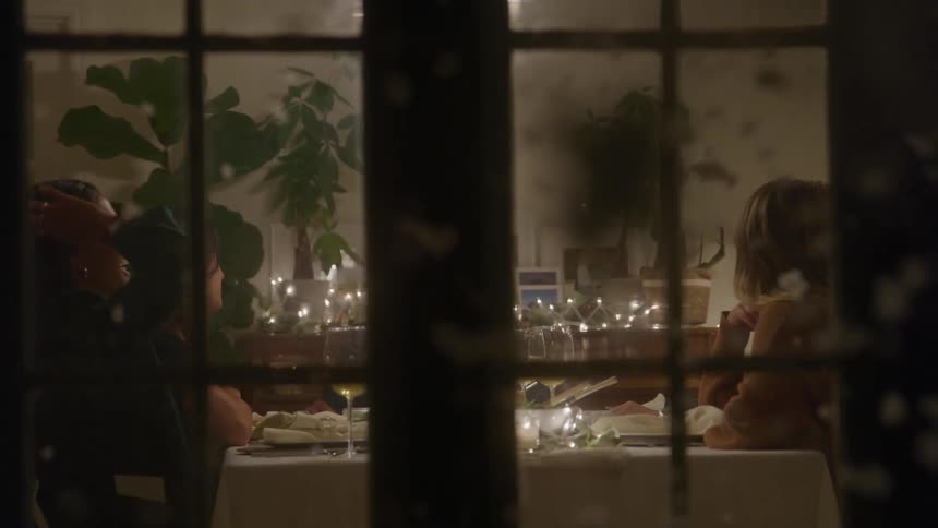 Based on the images from the video a group of people is sitting at a dining table seen through a large window the table is decorated with candles and holiday lights creating a warm and festive atmosphere