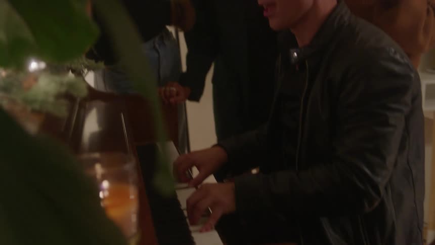 A group of friends having a house party as several women surround a young man playing the piano creating excitement in the room