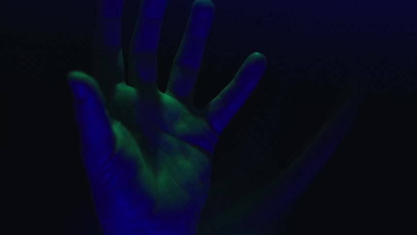 A single hand with the fingers curled is shown in a close view opening its palm toward the camera and rotating in a slow upward motion while being illuminated by shifting blue and green light against a dark background
