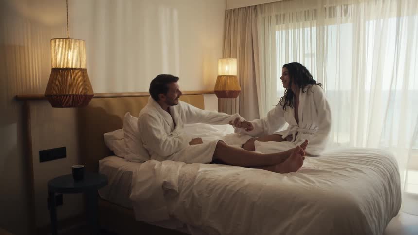 Couple in bathrobes sit playfully on a hotel bed