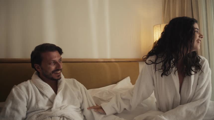 Couple wearing white bathrobes sitting on the bed smiling and looking to the side