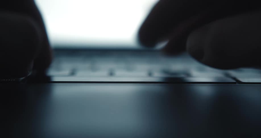 A close low angle view shows two hands typing on a light gray laptop keyboard in front of a bright screen