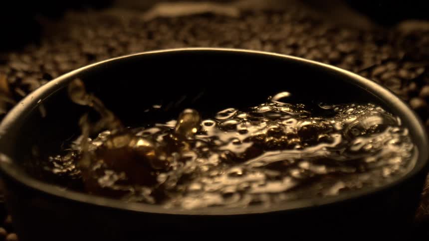 A close overhead view shows a dark coffee being poured into a black mug surrounded by coffee beans