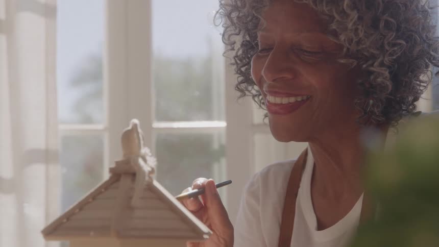 A woman with short gray curly hair is smiling and looking to the left as she sits in a sunlit room holding a small brush in her hand to paint a decorative wooden birdhouse