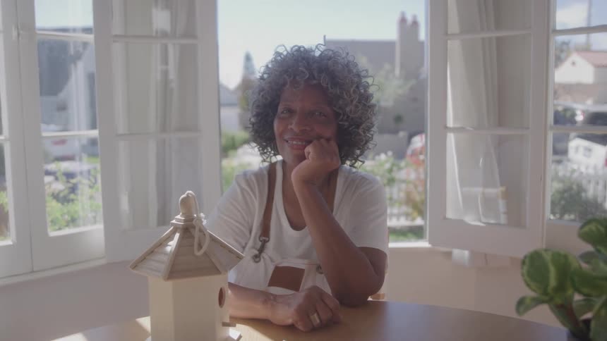 A smiling woman sitting at a table with a wooden bird house in front of her and an open window with a view to her neighborhood behind her