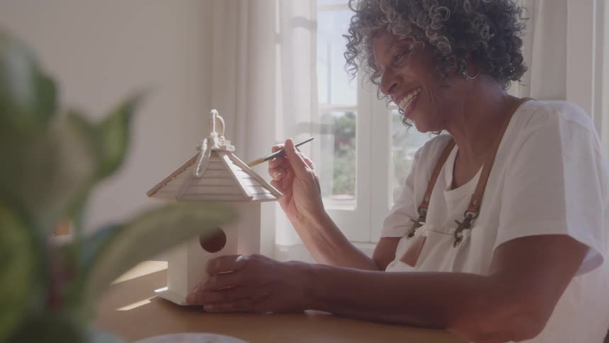 Based on the images from the video a woman with gray hair is seen smiling and looking down at something in her hand there is a plant in the foreground and sunlight is streaming in through a window behind her
