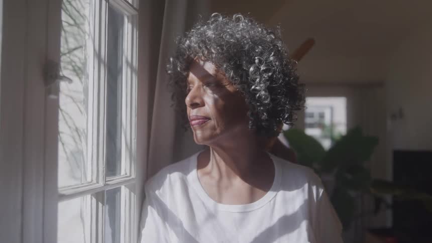 Woman looks thoughtfully out the window illuminated by sunlight
