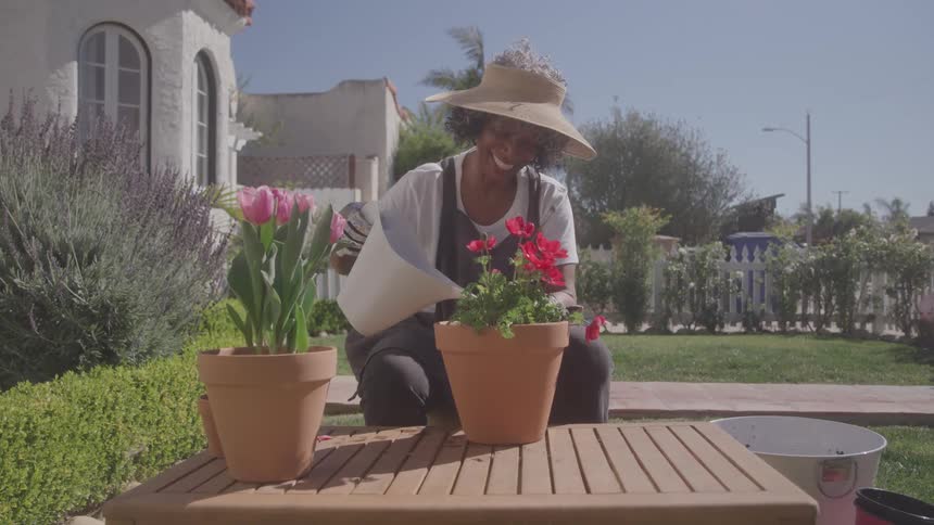 A smiling elderly woman wearing a hat and overalls crouched in a garden as she waters large flowers in pots