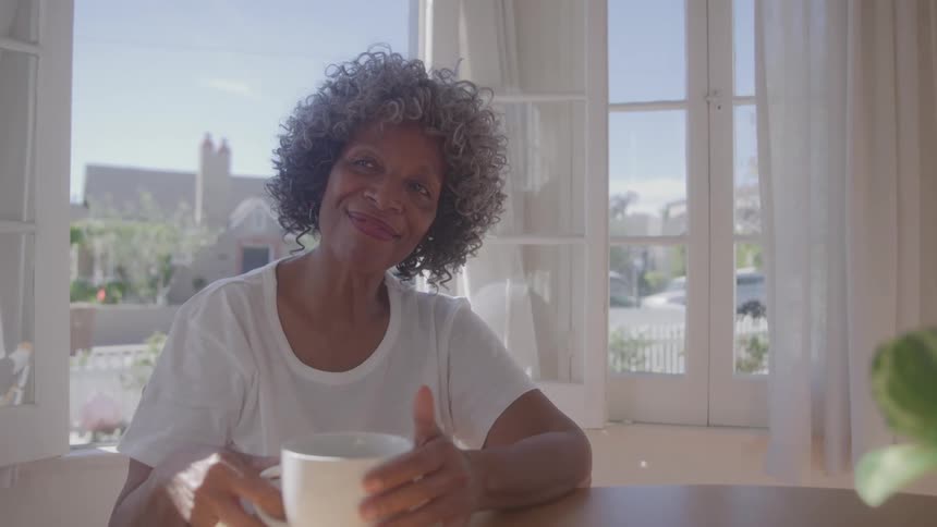 A woman with short gray curly hair is sitting at a wooden table in a sunlit room holding a white mug with both hands while looking directly at the camera smiling