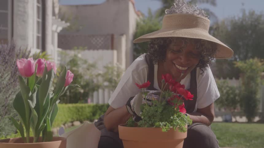 Smiling woman in a straw hat tends to flowers in pots in a sunny garden