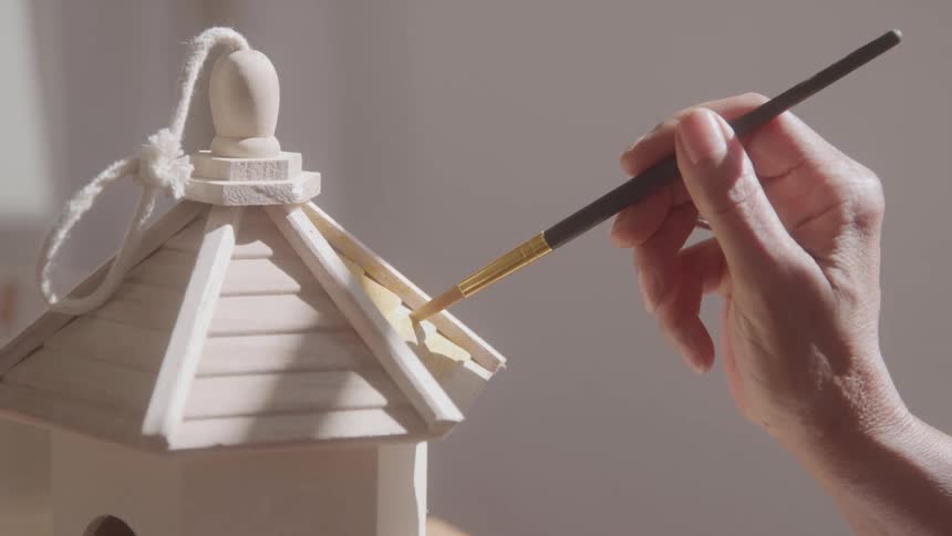 A womans hand using a thin brush to paint a wooden bird house