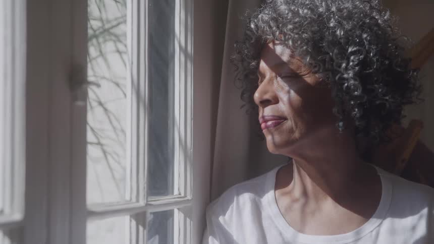 A woman stands next to a window an looks outside as a tree sways in the wind