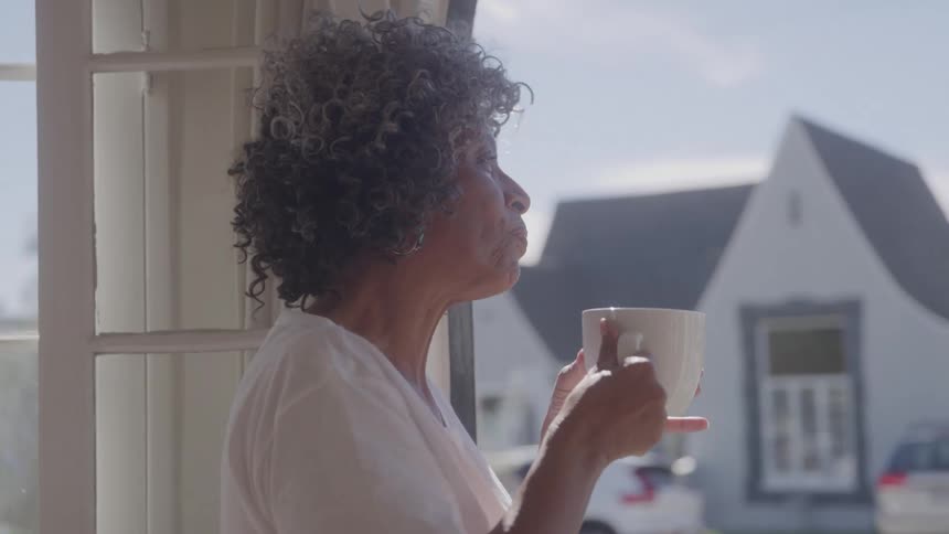 Elderly woman enjoys a hot drink while looking out the sunlit window