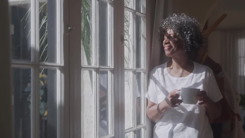 A woman with short gray curly hair is standing in a room with a window holding a white mug with both hands as she looks outside with a small smile on her face