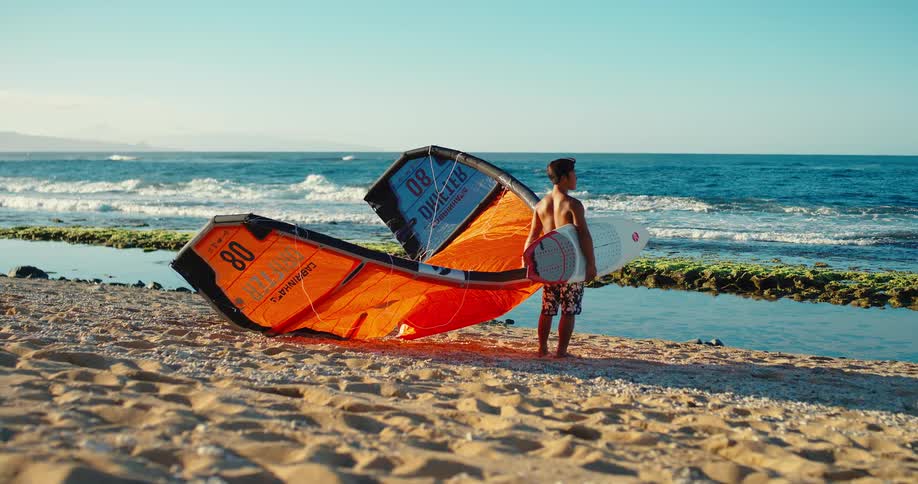 A man with a surfboard under his arm is standing barefoot on a sandy beach next to a large orange and blue kite looking out at the calm blue ocean