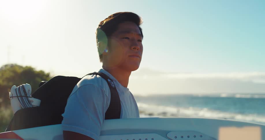 A man with short dark hair is shown from the chest up in a side view looking out at the ocean while standing on a grassy cliff and holding a white surfboard under his arm