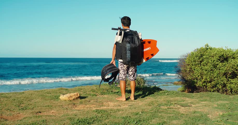 Based on the images from the video a man is seen from the back holding a surfboard and a backpack and looking out at the ocean the sky is bright blue and the waves are crashing on the shore in the background