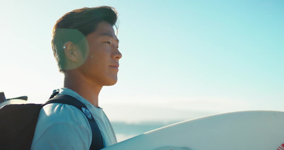 Young man wearing a backpack stands along the coast holding his surfboard