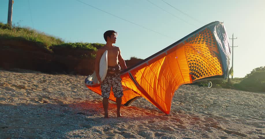 A man carries a surfboard and a kite sail on a sandy beach