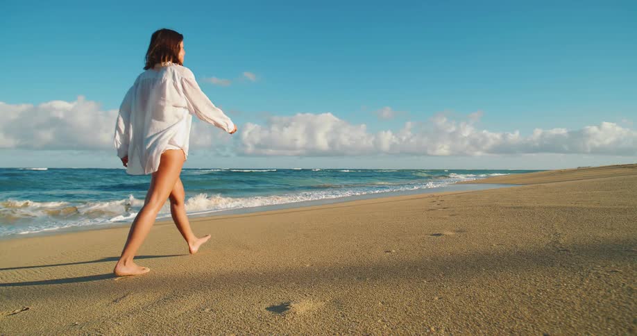 A woman with long brown hair wearing a loose white shirt and dark shorts is walking barefoot on a sandy beach at sunset with blue ocean waves approaching the shore