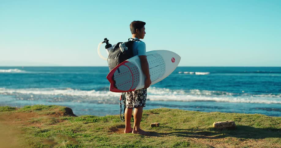 A man with dark hair wearing a gray t shirt patterned shorts and a dark backpack with a light colored hydration pack attached is standing barefoot on a grassy hill overlooking the ocean while holding a white surfboard under his left arm
