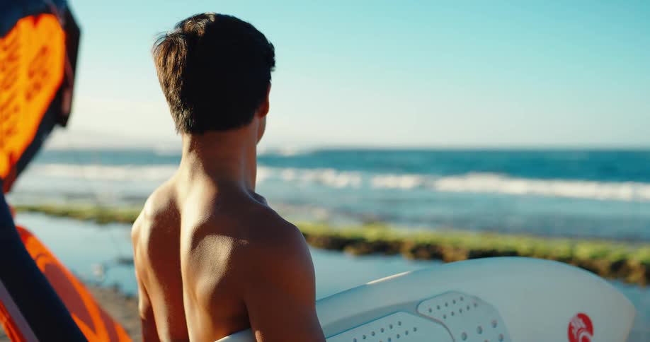 Man seen from behind holds a surfboard while looking at the sea