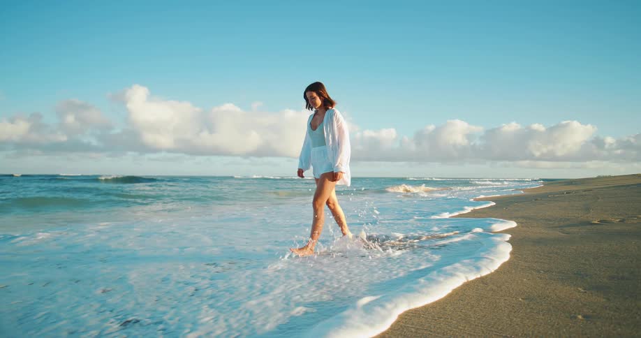 Young woman walks on the beach as waves roll into shore and over her bare feet as she walks in the water