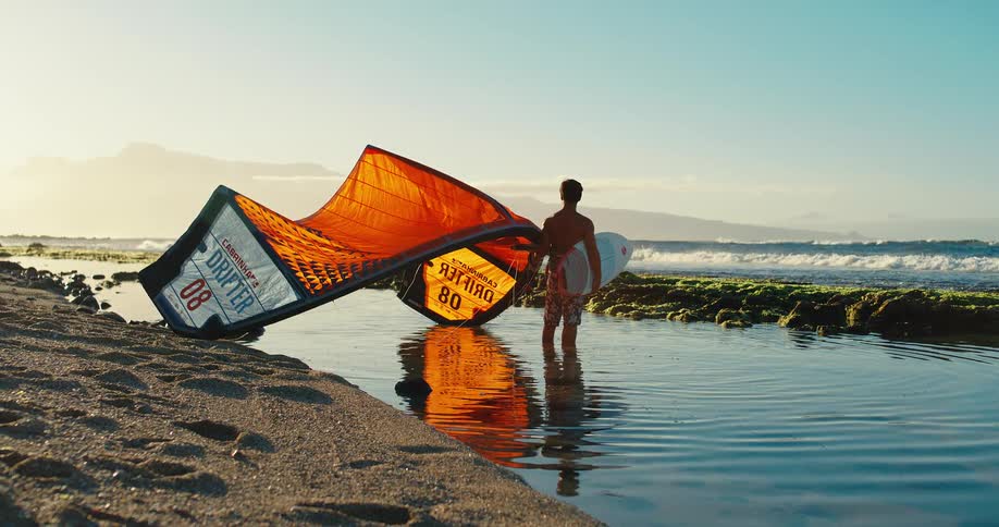 The man is holding his surfing board and kite while standing in the water