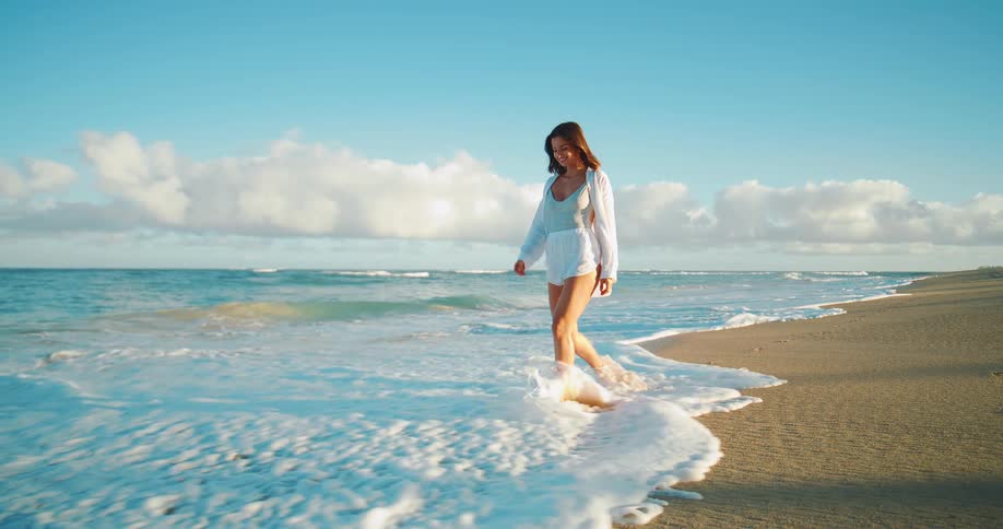Young woman walks on the beach as waves roll into shore and over her bare feet as she walks in the water