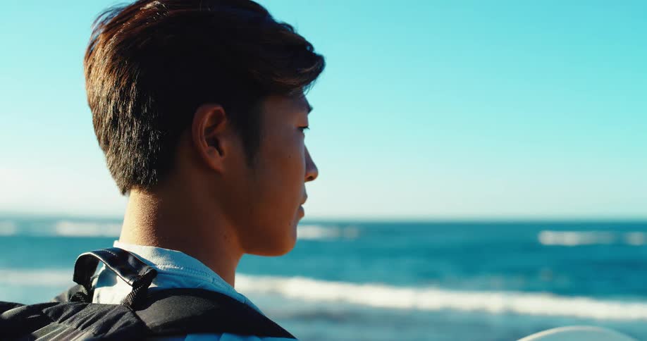 A man with short dark hair is shown in a side view looking out at the ocean with a neutral expression while standing on a beach with a white surfboard under his arm