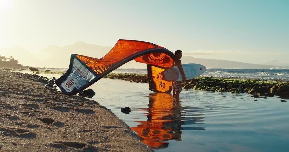 A young man stands looking at the ocean holding equipment for kite surfing