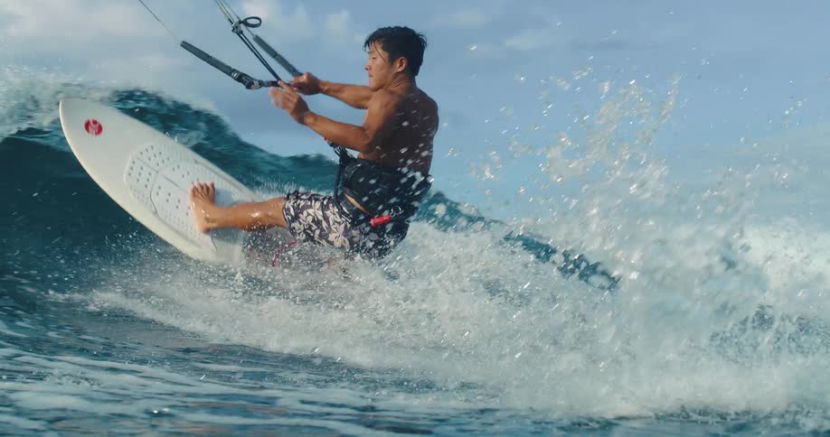A man wearing a black harness and patterned shorts is kiteboarding on a blue ocean wave creating a large white splash of water as he leans to the left while holding a control bar attached to a kite flying overhead
