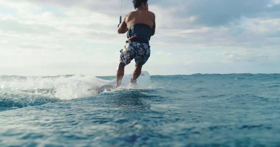 A view from the water shows a mans legs and the white surfboard he is riding as he glides across the surface with his body then coming into full view as he rides a wave with a kite control bar in his hands