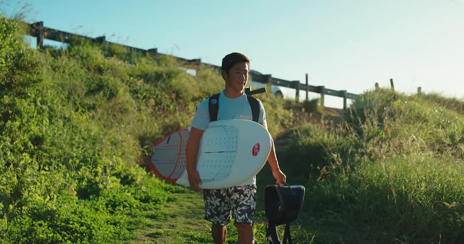 A young man wearing a light blue tshirt and floral shorts walks along a dirt path on a grassy hill carrying a white surfboard and a black bag while looking to his side