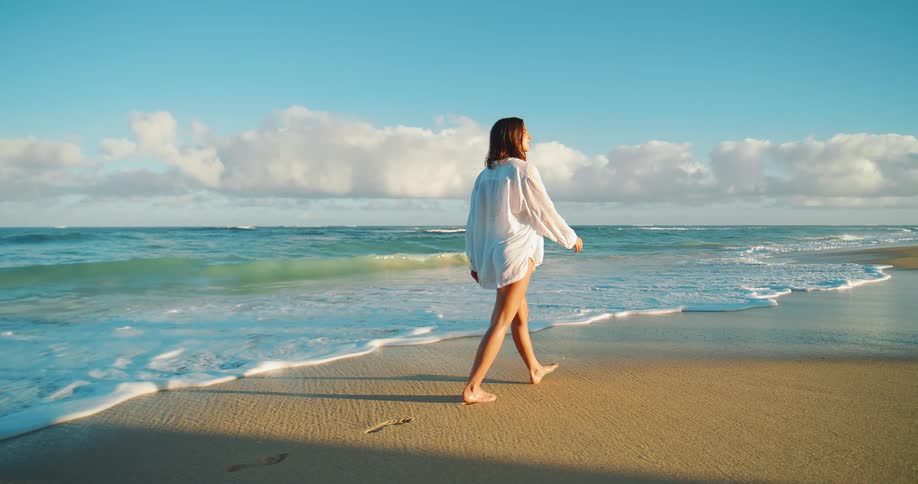 A woman with dark hair wearing a loose white shirt and dark shorts is walking barefoot on a sandy beach at sunset with blue ocean waves approaching the shore