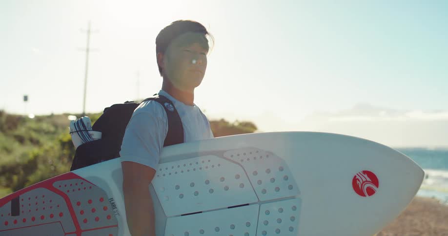 Young man wearing a backpack stands along the coast holding his surfboard
