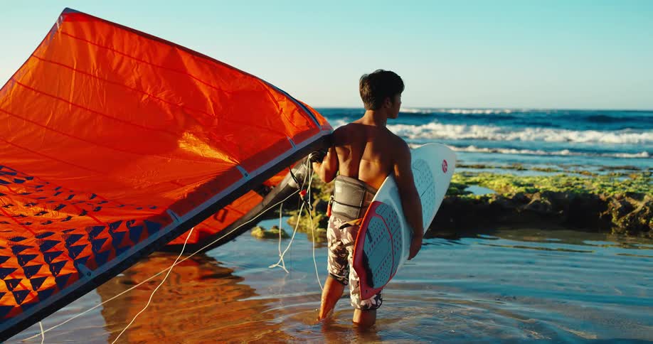 Surfer standing at the shore holding a surfboard and a kitesurf kite