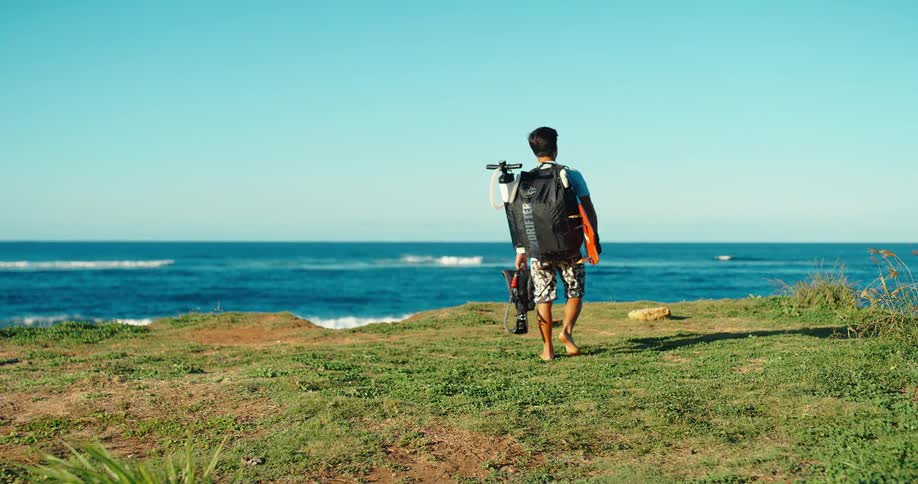 Based on the images from the video a man is seen from the back holding a surfboard and a backpack and looking out at the ocean the sky is bright blue and the waves are crashing on the shore in the background