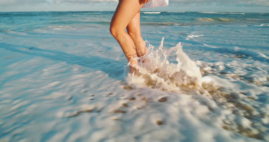Young woman walks on the beach as waves roll into shore and over her bare feet
