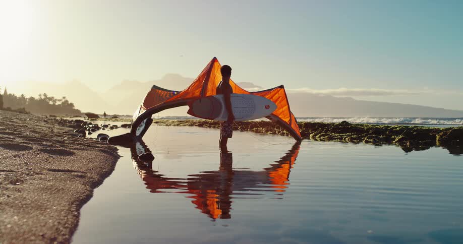 Person standing at the seashore holds a surfboard with an orange kite reflected in the water