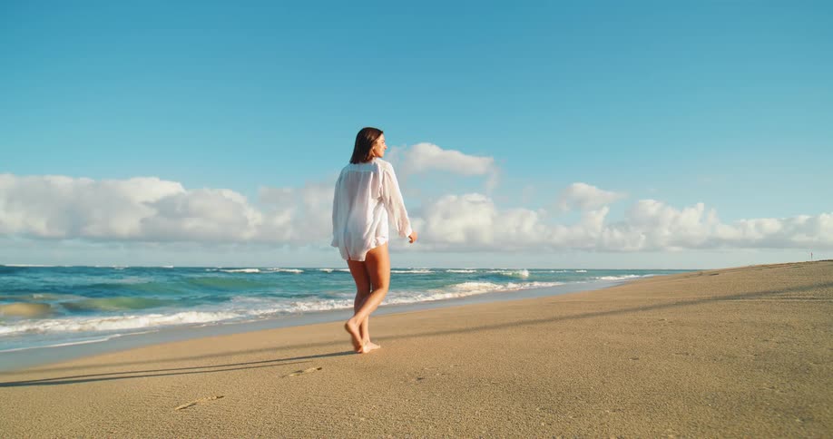 Based on the images from the video a woman wearing a white shirt is seen walking alone on a wide sandy beach the water is calm with small waves gently rolling onto the shore and the sky is a bright blue with scattered clouds