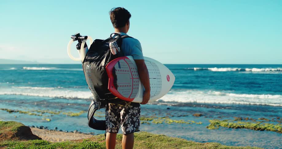 Young man wearing a backpack stands along the coast holding his surfboard as he watches the waves roll into shore