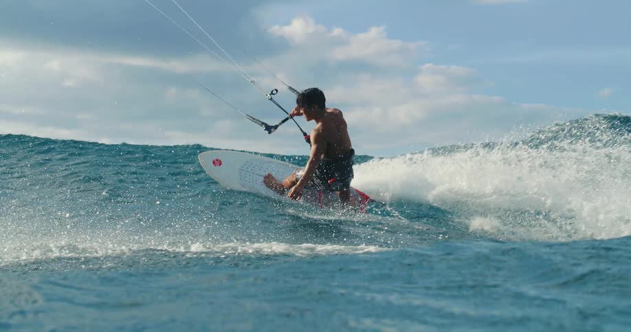 A man wearing patterned swim trunks and a harness is riding a white surfboard gripping the control bar of a kite and creating a large splash of white water as he rides a blue ocean wave