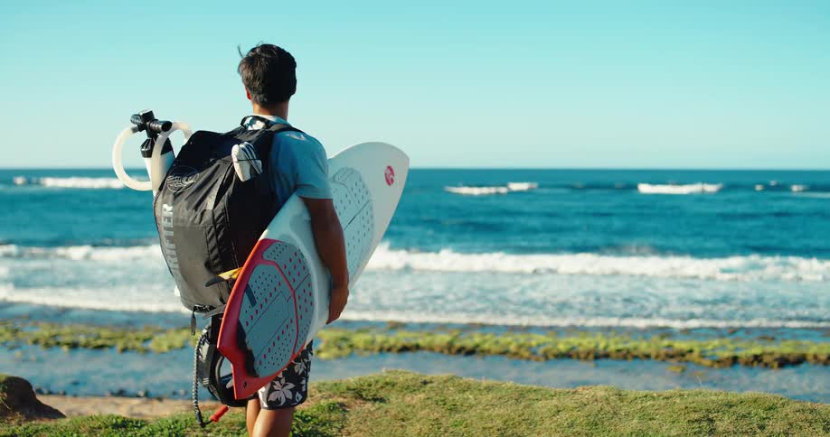 A man wearing a black backpack and a light blue shirt is standing on a grassy hill holding a white and red surfboard under his arm while looking out at the calm blue ocean