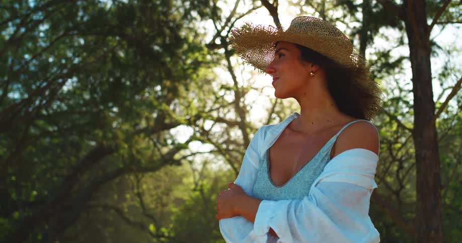 A woman wearing a large straw sun hat is standing in a sunlit forest with a white button down shirt hanging over a swimsuit as she turns her head to the left and smiles