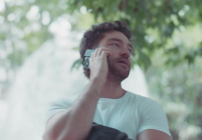 Man in a white tshirt talking on the phone in a treelined outdoor area