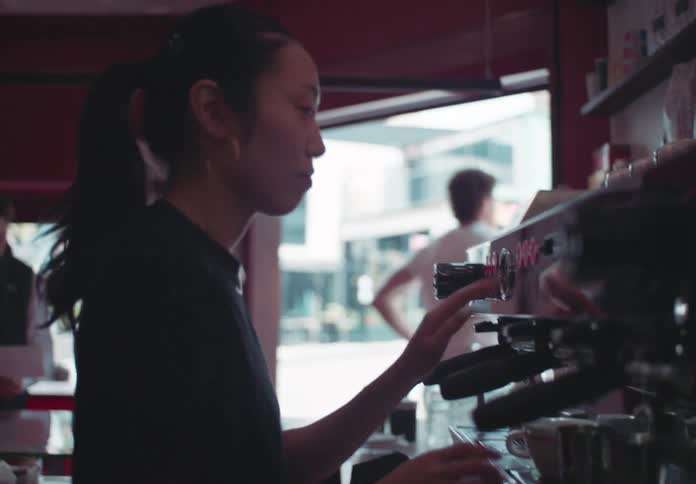 A barista is standing behind a counter in a coffee shop using a large espresso machine to make a drink while customers waits in the background near the counter