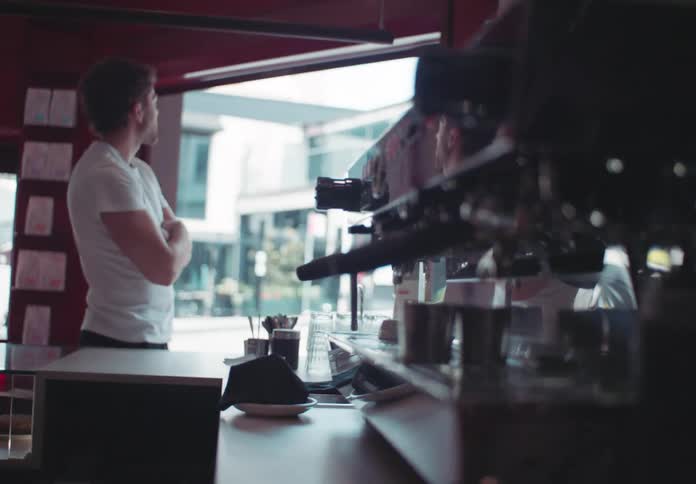 A customer in a white shirt and dark pants stands in front of the counter of a coffee shop with his arms crossed as he looks out a large front window at the street