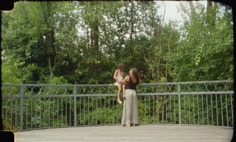 A woman and a young girl together with the girl sitting up on the safety rail of a lookout point to a forest and the mother holding her as they both talk and laugh