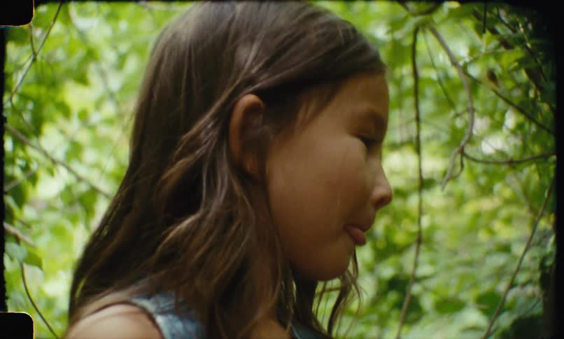 A young girl with long brown hair smiles and looks down with a blurry green foliage background
